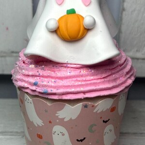 May include: A Halloween-themed cupcake with pink frosting and a decorative ghost topper holding an orange pumpkin. The cupcake liner features a repeating pattern of ghosts, bats, and stars, creating a festive and whimsical treat.