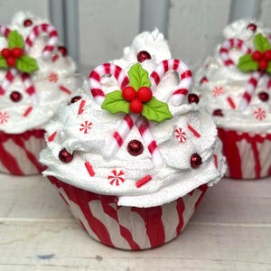 May include: Three decorative cupcakes with white frosting, red and white striped cupcake liners, and candy cane decorations. Each cupcake is topped with candy canes, red berries, and green holly leaves. Red sprinkles and glitter add sparkle.