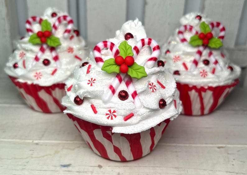 May include: Three decorative cupcakes with white frosting, red and white striped cupcake liners, and candy cane decorations. Each cupcake is topped with candy canes, red berries, and green holly leaves. Red sprinkles and glitter add sparkle.