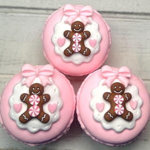 May include: Three pink macarons with gingerbread man decorations. Each macaron has a pink bow, a white scalloped edge, a brown gingerbread man with pink and white candy accents, and pink heart details.