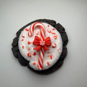 May include: A chocolate cookie topped with white frosting and red and white candy cane sprinkles. A red bow and candy cane are on top of the frosting.