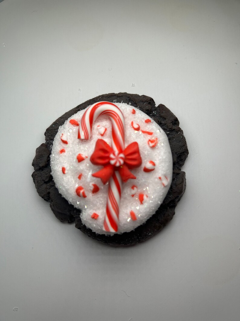 May include: A chocolate cookie topped with white frosting and red and white candy cane sprinkles. A red bow and candy cane are on top of the frosting.