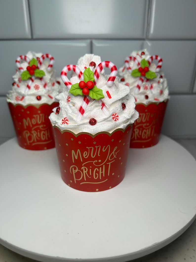 Pode incluir: Tr&ecirc;s forros de cupcake vermelhos e brancos com cobertura branca, bast&otilde;es de caramelo vermelhos e brancos e azevinho verde. Os forros de cupcake t&ecirc;m letras douradas que dizem "Merry & Bright".