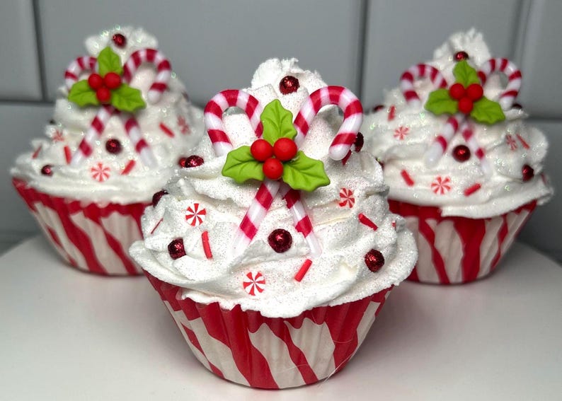 May include: Three festive cupcakes with white frosting, red and white striped candy canes, and red berry accents. The cupcakes sit in red and white striped paper cups, perfect for holiday celebrations.
