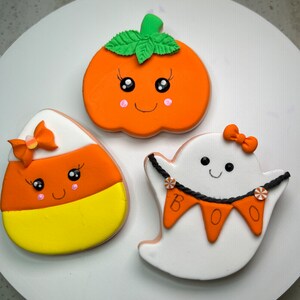 Pode incluir: Tr&ecirc;s biscoitos de a&ccedil;&uacute;car com tema de Halloween decorados com glac&ecirc; laranja, amarelo, branco e verde. Os biscoitos t&ecirc;m a forma de uma ab&oacute;bora, um fantasma e um milho doce. A ab&oacute;bora tem um caule e folhas verdes, e o fantasma tem uma corda de contas pretas e uma faixa que diz "BOO".