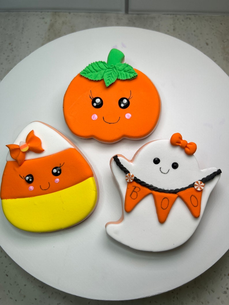 Pode incluir: Tr&ecirc;s biscoitos de a&ccedil;&uacute;car com tema de Halloween decorados com glac&ecirc; laranja, amarelo, branco e verde. Os biscoitos t&ecirc;m a forma de uma ab&oacute;bora, um fantasma e um milho doce. A ab&oacute;bora tem um caule e folhas verdes, e o fantasma tem uma corda de contas pretas e uma faixa que diz "BOO".