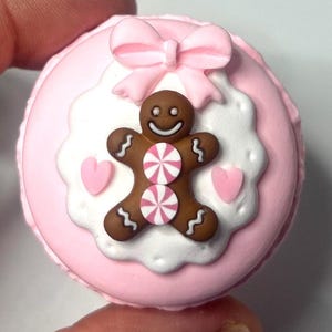 May include: A pink macaron with a gingerbread man design. The gingerbread man is brown with white icing details and two pink and white striped candies. A pink bow and two pink hearts complete the design.