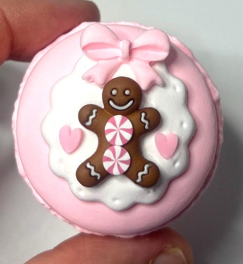 May include: A pink macaron with a gingerbread man design. The gingerbread man is brown with white icing details and two pink and white striped candies. A pink bow and two pink hearts complete the design.