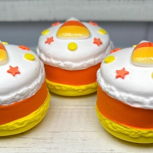 Puede incluir: Tres objetos decorativos con tem&aacute;tica de caramelo de ma&iacute;z. Cada uno tiene una parte superior blanca con estrellas naranjas y detalles de puntos amarillos, una base amarilla y una secci&oacute;n media naranja. Una pieza de caramelo de ma&iacute;z se encuentra encima. Los objetos est&aacute;n sobre una superficie de madera clara.