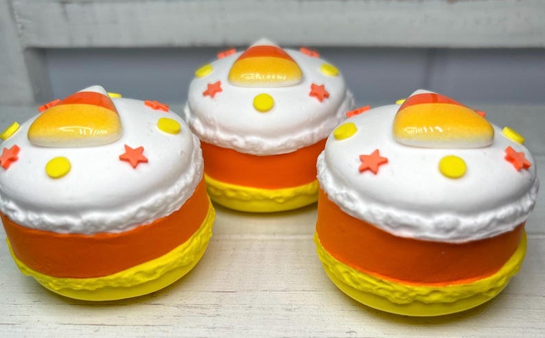 Puede incluir: Tres objetos decorativos con tem&aacute;tica de caramelo de ma&iacute;z. Cada uno tiene una parte superior blanca con estrellas naranjas y detalles de puntos amarillos, una base amarilla y una secci&oacute;n media naranja. Una pieza de caramelo de ma&iacute;z se encuentra encima. Los objetos est&aacute;n sobre una superficie de madera clara.