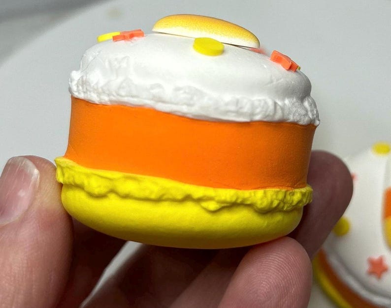 Puede incluir: Primer plano de una decoraci&oacute;n en forma de caramelo de ma&iacute;z. La decoraci&oacute;n tiene una parte superior blanca con chispas, un centro naranja y una parte inferior amarilla. Una peque&ntilde;a pieza amarilla y naranja est&aacute; en la parte superior.