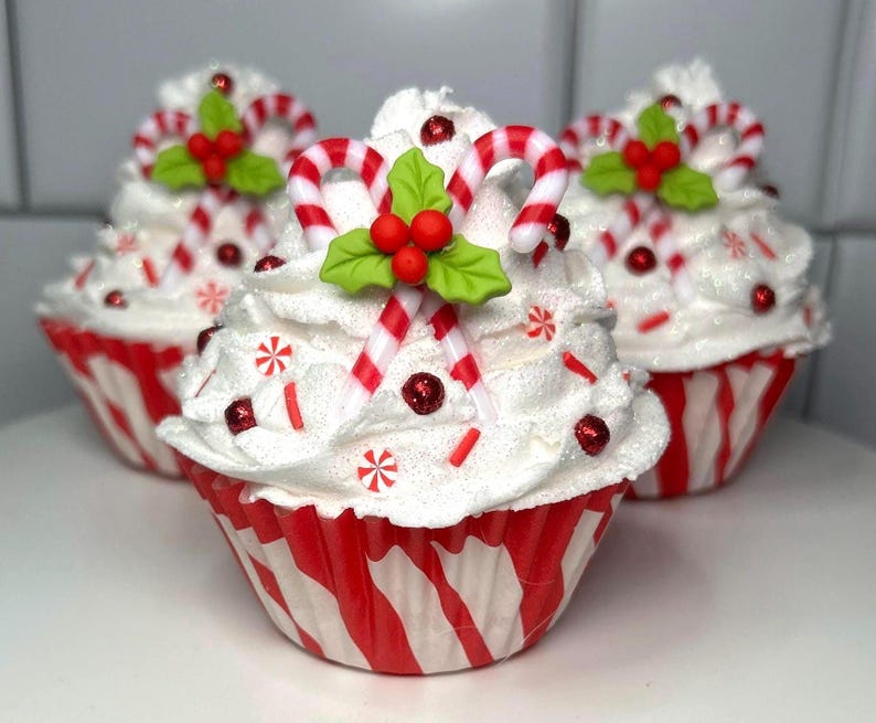 May include: Three festive cupcakes with white frosting, red and white striped wrappers, and candy cane decorations. Each cupcake is topped with a candy cane, holly leaves, and red berry accents, perfect for the holiday season.