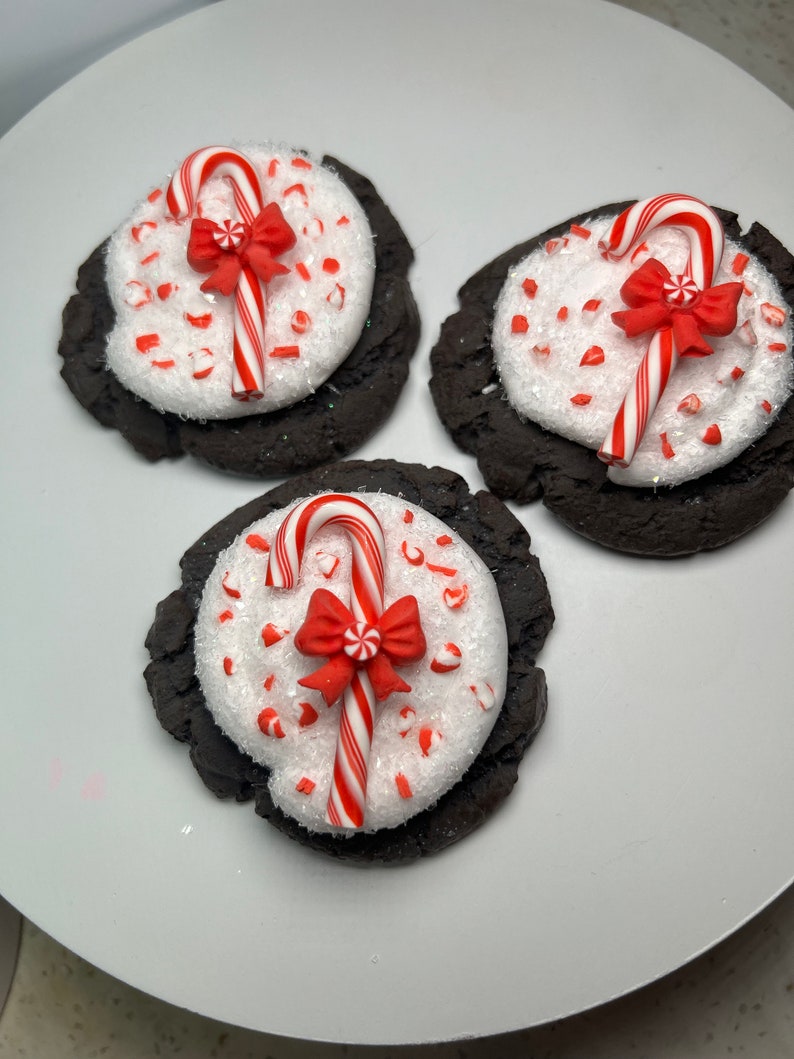 May include: Three chocolate cookies decorated with white frosting, red and white candy canes, and red bows. The cookies are on a white plate.