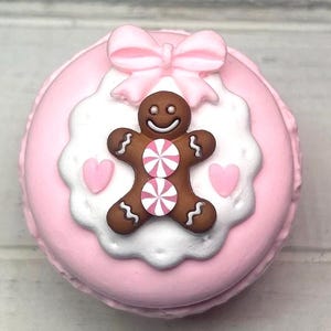 May include: A pink macaron decorated with a gingerbread man, pink bow, and hearts. The gingerbread man has two candy accents and a smiling face. The macaron is a light pink color with a white and pink frosting design.