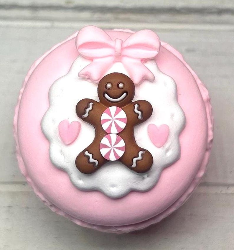 May include: A pink macaron decorated with a gingerbread man, pink bow, and hearts. The gingerbread man has two candy accents and a smiling face. The macaron is a light pink color with a white and pink frosting design.