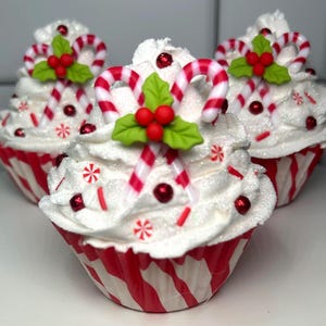 May include: Three festive cupcakes with white frosting, red and white striped wrappers, and candy cane decorations. Each cupcake is topped with a candy cane, red berries, green leaves, and red sprinkles, creating a holiday theme.
