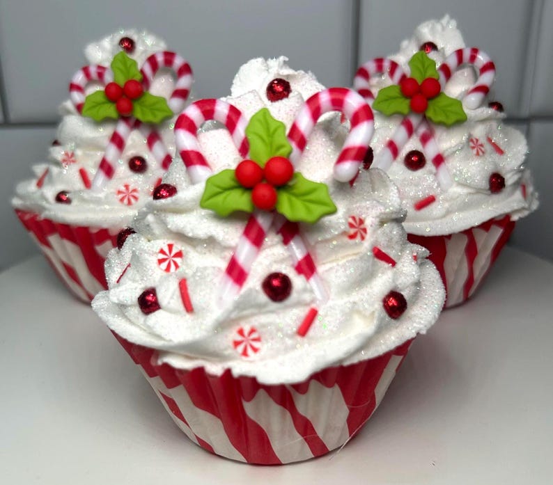 May include: Three festive cupcakes with white frosting, red and white striped wrappers, and candy cane decorations. Each cupcake is topped with a candy cane, red berries, green leaves, and red sprinkles, creating a holiday theme.