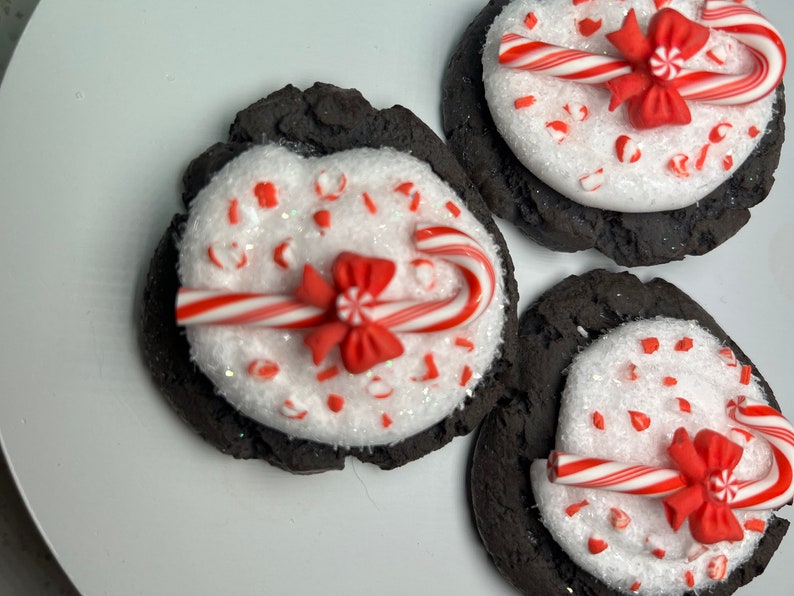 May include: Three chocolate cookies decorated with white frosting, red and white candy canes, and red bows. The cookies are on a white plate.