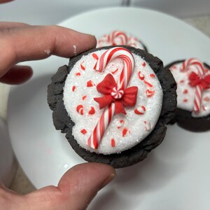 May include: Close-up of a chocolate cookie topped with white frosting, red and white candy cane sprinkles, a red bow, and a candy cane.