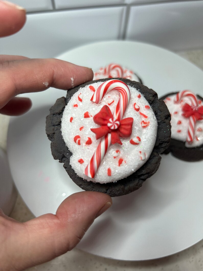 May include: Close-up of a chocolate cookie topped with white frosting, red and white candy cane sprinkles, a red bow, and a candy cane.