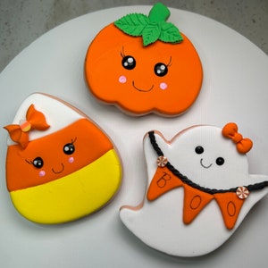 Pode incluir: Tr&ecirc;s biscoitos de a&ccedil;&uacute;car com tema de Halloween decorados com glac&ecirc;. Um biscoito de ab&oacute;bora com uma folha verde, um biscoito de milho doce com um la&ccedil;o rosa e um biscoito de fantasma com uma faixa de "BOO".