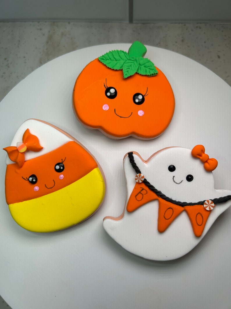 Pode incluir: Tr&ecirc;s biscoitos de a&ccedil;&uacute;car com tema de Halloween decorados com glac&ecirc;. Um biscoito de ab&oacute;bora com uma folha verde, um biscoito de milho doce com um la&ccedil;o rosa e um biscoito de fantasma com uma faixa de "BOO".