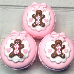 May include: Three pink macarons with gingerbread man decorations. Each macaron has a bow on top, a white circular base, and a brown gingerbread man with pink and white candy accents. Two pink hearts are on the white base.