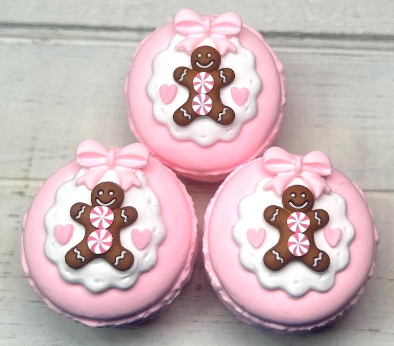 May include: Three pink macarons with gingerbread man decorations. Each macaron has a bow on top, a white circular base, and a brown gingerbread man with pink and white candy accents. Two pink hearts are on the white base.