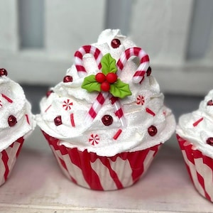 May include: Three festive cupcakes with white frosting, red and white striped wrappers, and candy cane decorations. Each cupcake is topped with a candy cane, holly leaves, and red berry accents, perfect for the holiday season.