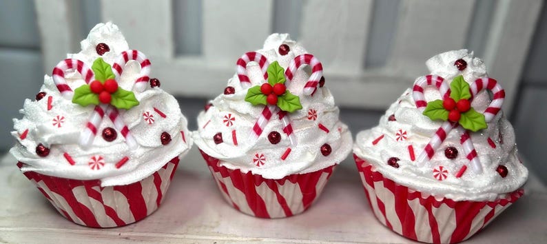 May include: Three festive cupcakes with white frosting, red and white striped wrappers, and candy cane decorations. Each cupcake is topped with a candy cane, holly leaves, and red berry accents, perfect for the holiday season.