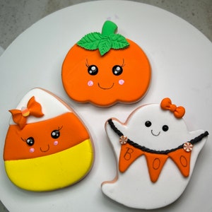 Pode incluir: Tr&ecirc;s biscoitos de a&ccedil;&uacute;car tem&aacute;ticos de Halloween decorados com glac&ecirc; colorido. Os biscoitos s&atilde;o em forma de ab&oacute;bora, fantasma e milho doce. A ab&oacute;bora tem um caule verde e um rosto sorridente com bochechas rosadas. O fantasma tem um corpo branco com uma faixa preta que diz "BOO" e um la&ccedil;o rosa. O milho doce tem um design amarelo, laranja e branco com um rosto sorridente e um la&ccedil;o rosa.