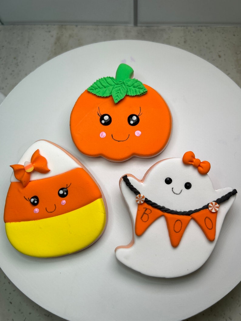 Pode incluir: Tr&ecirc;s biscoitos de a&ccedil;&uacute;car tem&aacute;ticos de Halloween decorados com glac&ecirc; colorido. Os biscoitos s&atilde;o em forma de ab&oacute;bora, fantasma e milho doce. A ab&oacute;bora tem um caule verde e um rosto sorridente com bochechas rosadas. O fantasma tem um corpo branco com uma faixa preta que diz "BOO" e um la&ccedil;o rosa. O milho doce tem um design amarelo, laranja e branco com um rosto sorridente e um la&ccedil;o rosa.
