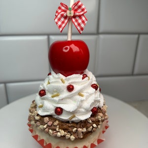 以下が含まれることがあります： 白と赤のカップケーキで、白いフロスティング、赤いスプリンクル、そして赤いキャンディーアップルがトッピングされています。キャンディーアップルには、白の棒に赤と白のチェック柄のリボンが付いています。