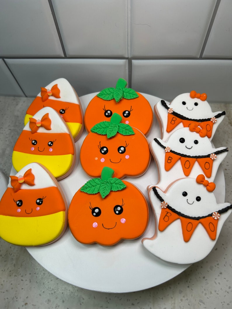 Pode incluir: Uma cole&ccedil;&atilde;o de nove biscoitos de a&ccedil;&uacute;car com tema de Halloween decorados com glac&ecirc; laranja, amarelo, branco e verde. Os biscoitos apresentam ab&oacute;boras, fantasmas e milho doce com carinhas fofas e la&ccedil;os.
