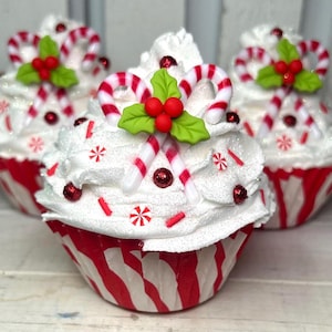 May include: Three decorative cupcakes with white frosting, red and white striped cupcake liners, and holiday-themed decorations. Each cupcake is topped with candy canes, red berries, and green holly leaves. Red sprinkles and glitter add festive detail.