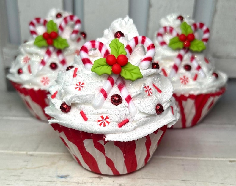 May include: Three decorative cupcakes with white frosting, red and white striped cupcake liners, and holiday-themed decorations. Each cupcake is topped with candy canes, red berries, and green holly leaves. Red sprinkles and glitter add festive detail.