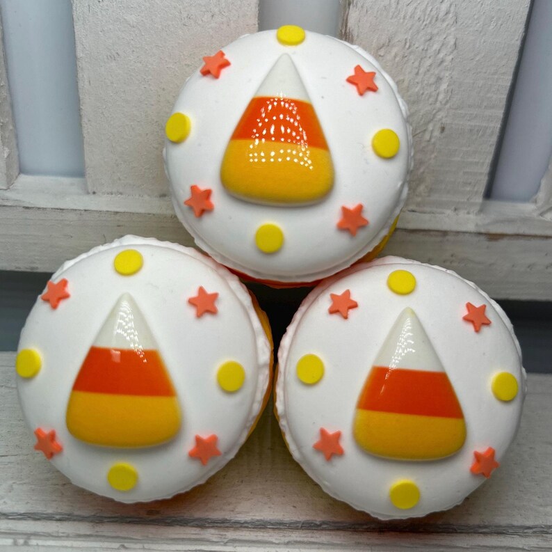 Puede incluir: Tres macarons blancos decorados con dise&ntilde;os de caramelos de ma&iacute;z. Cada macaron tiene un caramelo de ma&iacute;z en el centro, rodeado de estrellas naranjas y puntos amarillos. Los macarons est&aacute;n apilados.