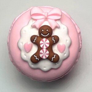 May include: A pink macaron decorated with a gingerbread man, pink bow, and white and pink accents. The gingerbread man has a smiling face and two peppermint candies on its body. Two pink hearts are on either side of the gingerbread man.