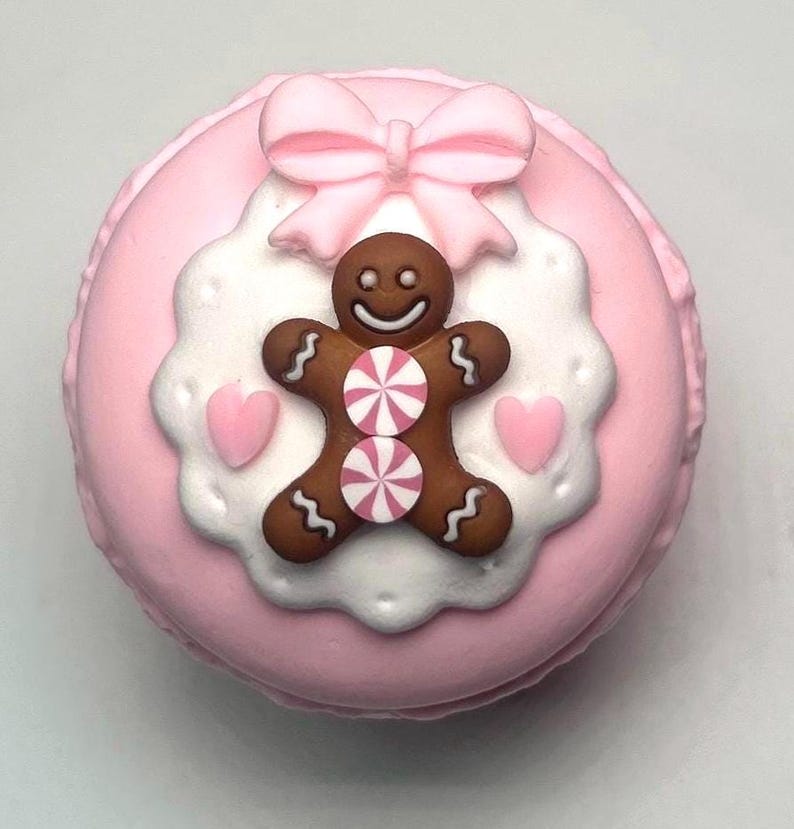 May include: A pink macaron decorated with a gingerbread man, pink bow, and white and pink accents. The gingerbread man has a smiling face and two peppermint candies on its body. Two pink hearts are on either side of the gingerbread man.