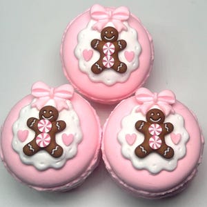 May include: Three pink macarons with gingerbread man decorations. Each macaron has a pink bow, a white scalloped edge, a brown gingerbread man with a pink and white candy center, and pink heart accents.
