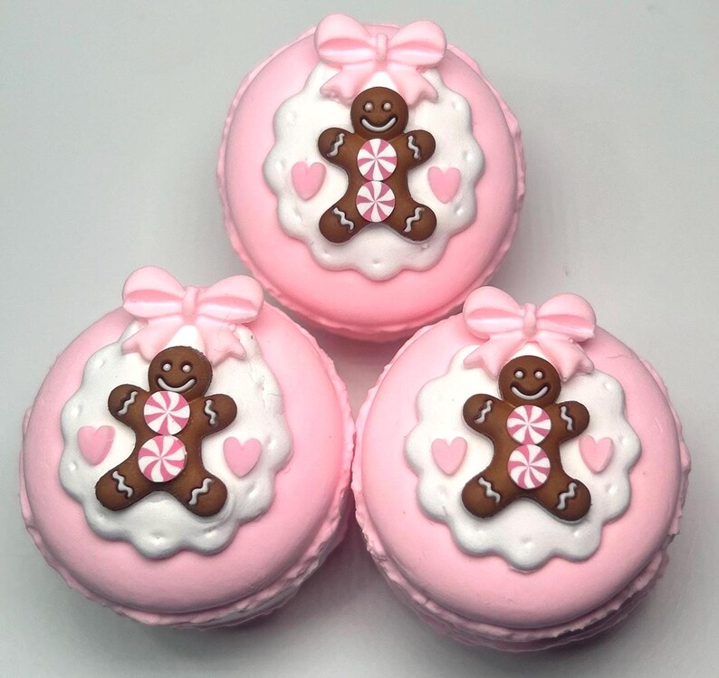 May include: Three pink macarons with gingerbread man decorations. Each macaron has a pink bow, a white scalloped edge, a brown gingerbread man with a pink and white candy center, and pink heart accents.