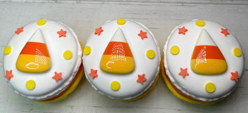 Puede incluir: Tres dulces redondos glaseados de blanco con dise&ntilde;os de caramelos de ma&iacute;z. Cada uno tiene un caramelo de ma&iacute;z en el centro, rodeado de estrellas naranjas y puntos amarillos. Los lados est&aacute;n en capas de amarillo y naranja.