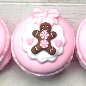 May include: Three pink macarons with gingerbread man decorations. Each macaron has a gingerbread man with pink and white striped candy buttons, a white scalloped border, pink hearts, and a pink bow.