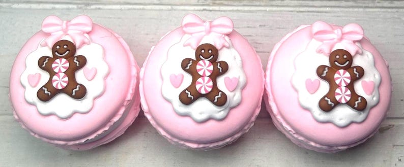 May include: Three pink macarons with gingerbread man decorations. Each macaron has a gingerbread man with pink and white striped candy buttons, a white scalloped border, pink hearts, and a pink bow.