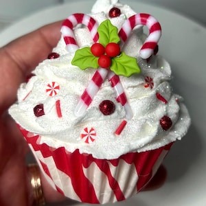 May include: A festive cupcake with white frosting, decorated with red and white candy canes, holly leaves, and red sprinkles. The cupcake sits in a red and white striped paper cup, perfect for holiday celebrations.
