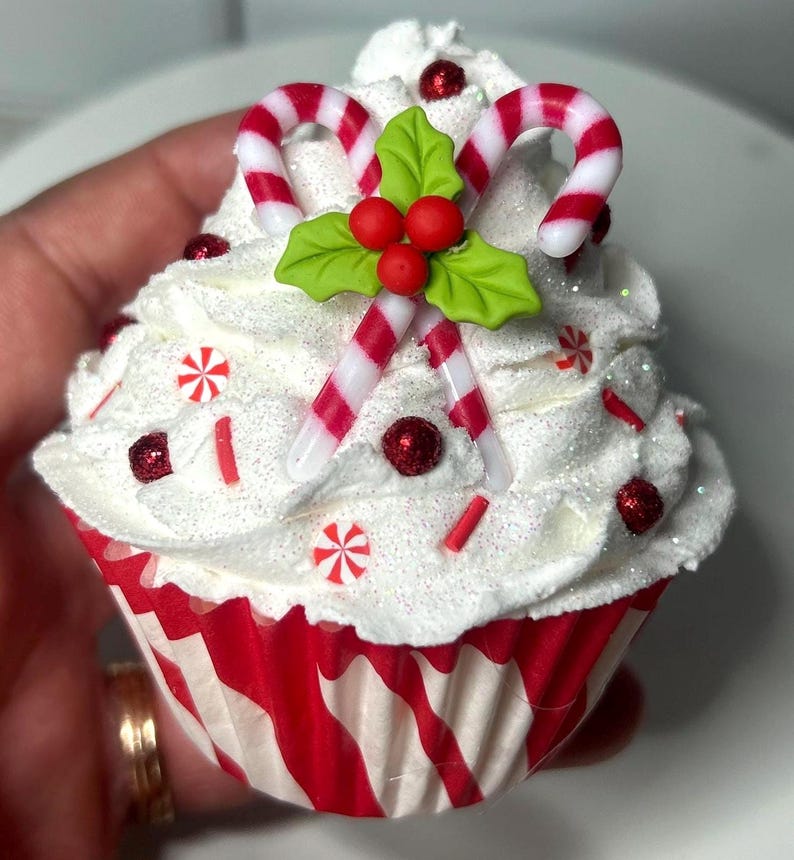 May include: A festive cupcake with white frosting, decorated with red and white candy canes, holly leaves, and red sprinkles. The cupcake sits in a red and white striped paper cup, perfect for holiday celebrations.