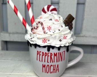 May include: A white mug with the words "Peppermint Mocha" in red, topped with whipped cream, candy, and chocolate. Two red and white striped straws and a candy cane are placed in the whipped cream. The mug has a black drip design.