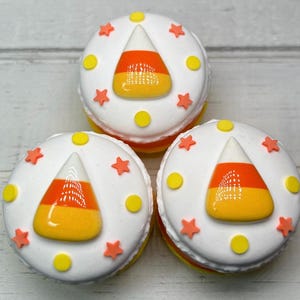 Puede incluir: Tres dulces redondos glaseados de blanco con dise&ntilde;os de caramelos de ma&iacute;z. Cada dulce tiene un caramelo de ma&iacute;z en el centro, rodeado de estrellas naranjas y puntos amarillos. Los dulces est&aacute;n apilados.