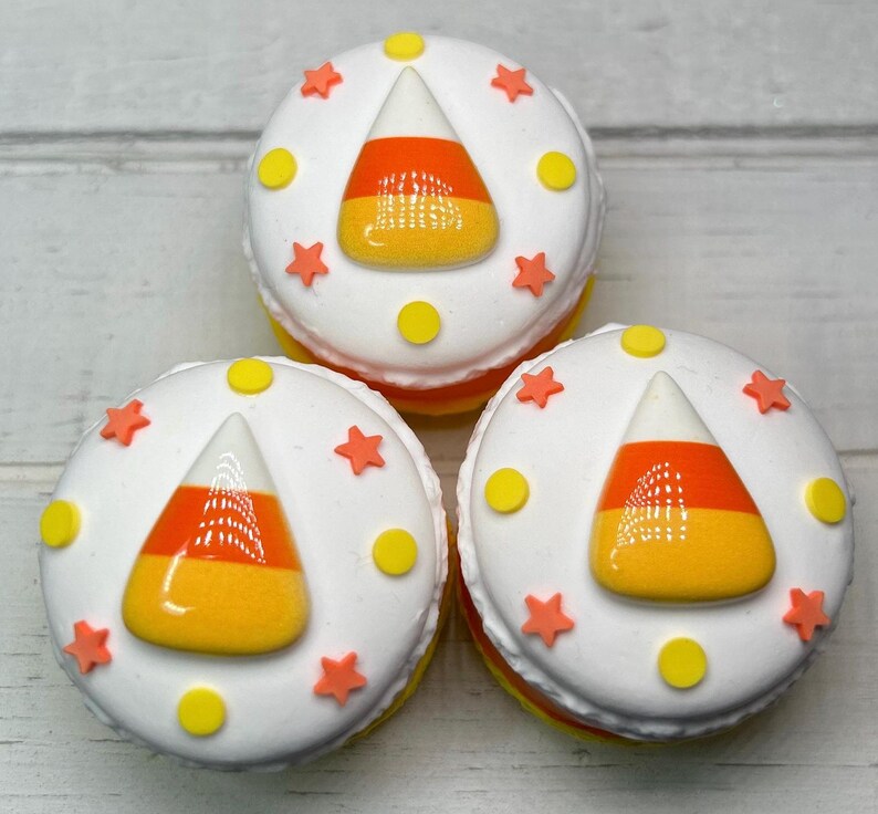 Puede incluir: Tres dulces redondos glaseados de blanco con dise&ntilde;os de caramelos de ma&iacute;z. Cada dulce tiene un caramelo de ma&iacute;z en el centro, rodeado de estrellas naranjas y puntos amarillos. Los dulces est&aacute;n apilados.