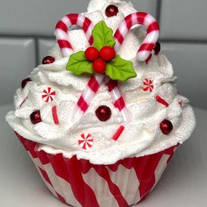 May include: A festive cupcake with white frosting, red and white candy canes, and holly berries. The cupcake is decorated with red sprinkles and candies, and sits in a red and white striped paper cup. A holiday-themed treat.